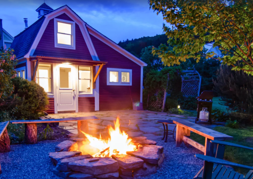 A cozy outdoor scene featuring a fire pit surrounded by benches, with a charming house and trees in the background at dusk.