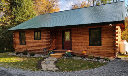 A cozy log cabin with a green metal roof, surrounded by trees and autumn foliage, featuring a stone pathway and flower beds.