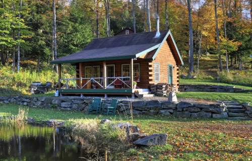 A cozy log cabin surrounded by colorful autumn trees, with a porch and a small pond nearby.