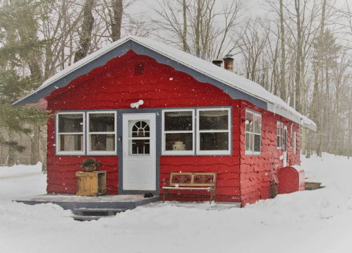 A bright red cabin surrounded by snow, with a white door and windows, nestled among leafless trees in winter.
