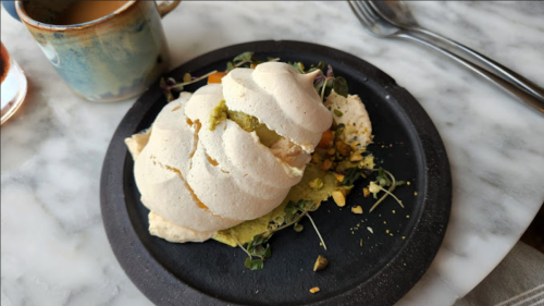 A meringue dessert topped with greens and nuts, served on a black plate beside a cup of coffee.