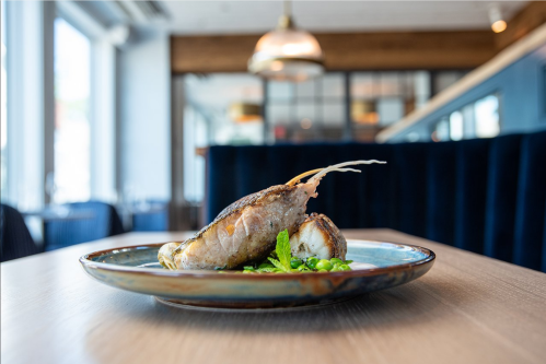 A beautifully plated fish dish garnished with greens, set on a wooden table in a modern restaurant interior.