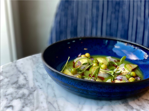 A vibrant dish with green olives, herbs, and sliced vegetables served in a deep blue bowl on a marble table.