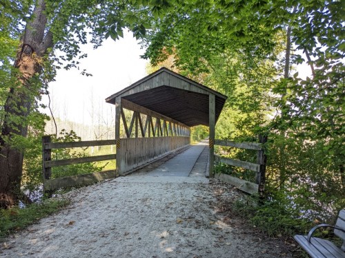A covered wooden bridge surrounded by trees, leading over a path by a serene body of water.