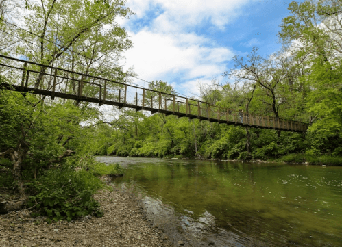 A rustic metal bridge spans a serene river, surrounded by lush green trees and a bright blue sky.
