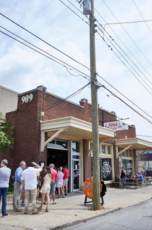 A line of people waiting outside a brick building with a sign, under a cloudy sky and power lines overhead.