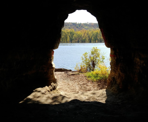 View from a cave opening overlooking a river and trees, with sunlight illuminating the entrance.