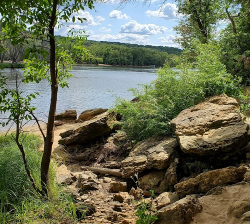 A serene riverside scene with rocks, greenery, and trees under a blue sky with fluffy clouds.
