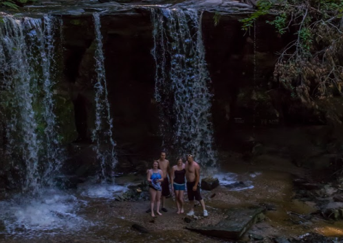 Four people stand in front of a waterfall, enjoying the natural scenery in a lush, green environment.