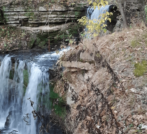 A serene waterfall cascades over rocky cliffs, surrounded by autumn foliage and a peaceful natural setting.