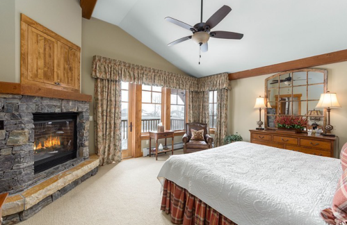 Cozy bedroom with a stone fireplace, large windows, and a patterned bedspread, featuring warm lighting and rustic decor.