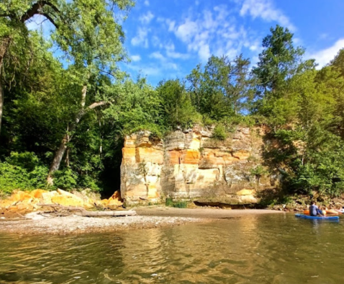 A serene river scene featuring a rocky cliff, lush greenery, and a clear blue sky, with a person kayaking nearby.