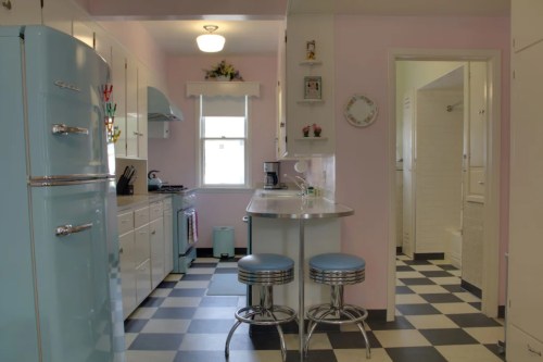 A retro-style kitchen with pastel blue appliances, checkered floor, and a small dining area. A bathroom is visible in the background.