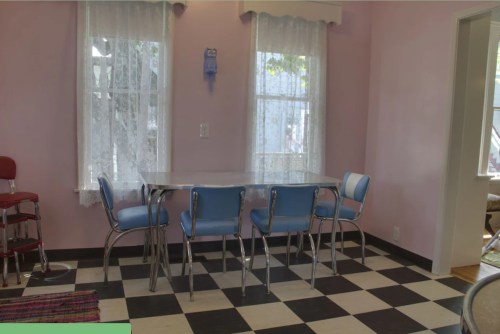 A pastel pink dining area with a checkered floor, featuring a table and blue chairs near large windows with white curtains.