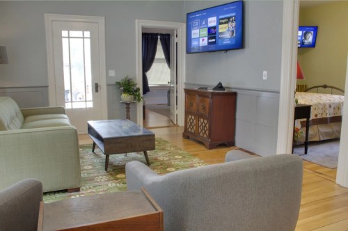 A cozy living room with a green sofa, coffee table, and a TV on the wall, leading to a bedroom and a door to the outside.