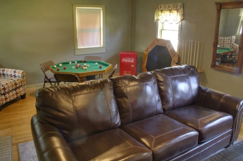 A cozy living room featuring a brown leather couch, a pool table, and a Coca-Cola vending machine in the background.
