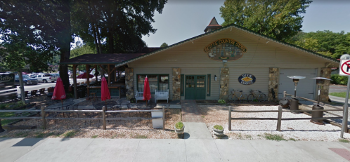 A cozy café with outdoor seating, red umbrellas, and a rustic stone exterior surrounded by trees.