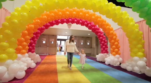 A woman and a young girl walk under a colorful balloon arch in a vibrant, rainbow-themed hallway.