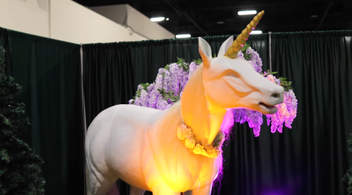 A life-sized white unicorn statue adorned with flowers and a golden horn, set against a dark backdrop.