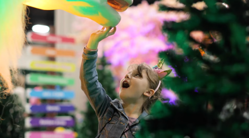 A young girl in a denim jacket reaches up in awe towards a colorful unicorn decoration amidst festive trees.