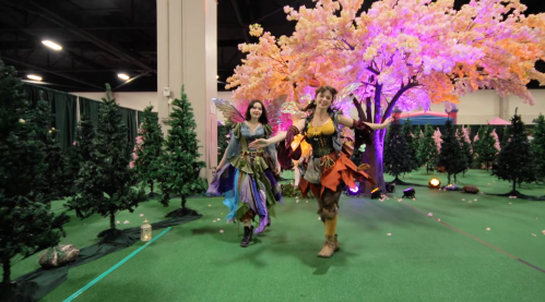 Two women in colorful fairy costumes walk joyfully through a whimsical forest setting with trees and pink lights.