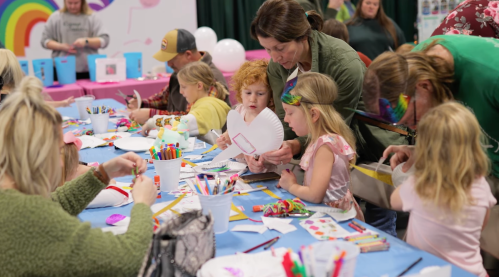 A group of children and adults engage in a craft activity at a colorful table, surrounded by art supplies and decorations.