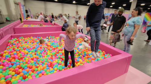Children play in a colorful ball pit at a lively event, surrounded by families and vibrant decorations.