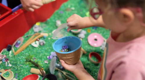 A child pours colorful gems from a container into a small blue pot while playing on a grassy surface.