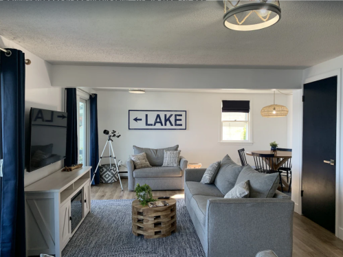 Cozy living room with gray furniture, lake-themed decor, and a dining area, featuring large windows and blue accents.