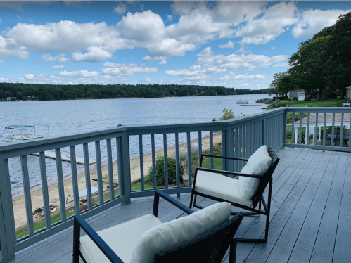 A serene lakeside view from a deck with two chairs, featuring a sandy beach and cloudy blue sky.