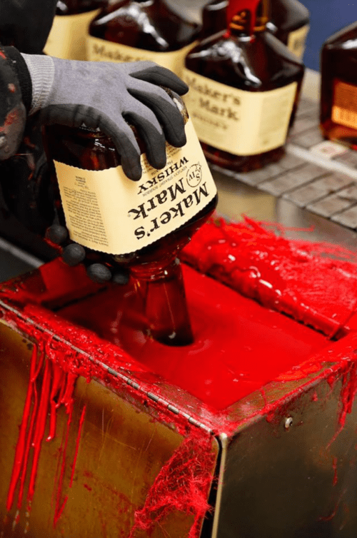 A gloved hand pours a bottle of whiskey into a container filled with red liquid, creating a striking visual contrast.