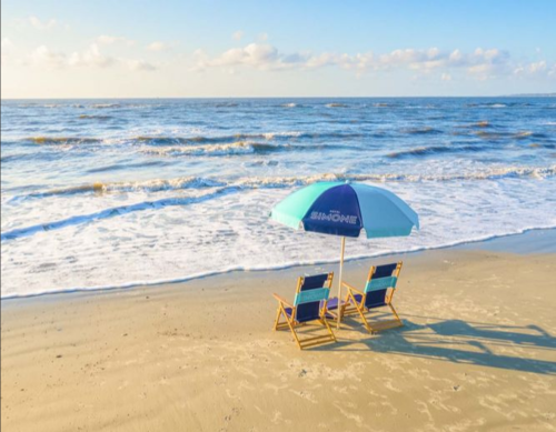 Two beach chairs under a blue and white umbrella on a sandy shore, with gentle waves lapping at the beach.