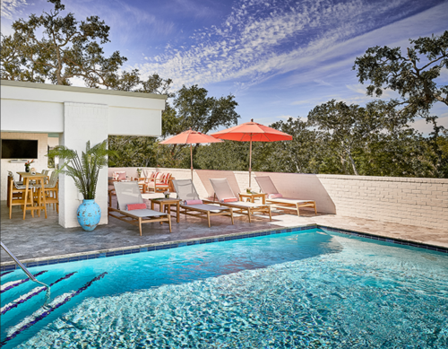 A sunny poolside scene with lounge chairs, umbrellas, and a vibrant blue vase, surrounded by trees and a clear sky.