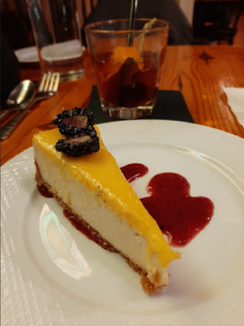 A slice of cheesecake topped with berries, served with raspberry sauce, alongside a glass of cocktail with an orange twist.