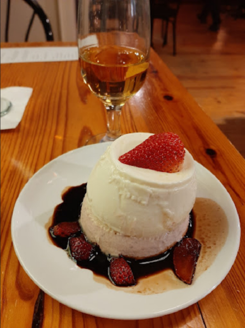 A creamy dessert topped with a strawberry, served on a plate with chocolate sauce and accompanied by a glass of amber drink.