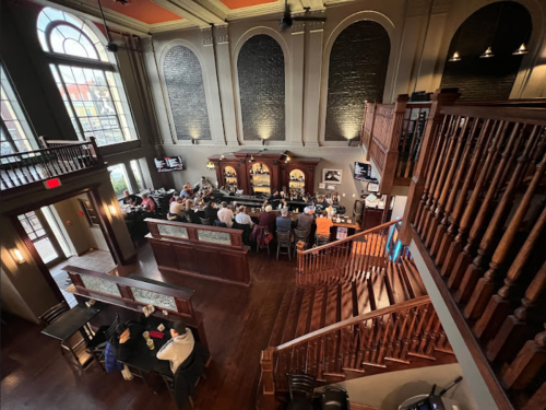 A spacious bar with a wooden staircase, high ceilings, and patrons seated at the counter and tables.
