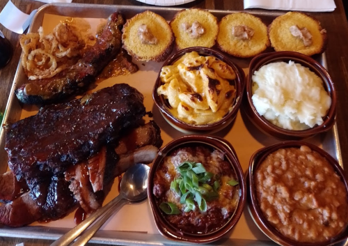 A metal tray filled with barbecue ribs, brisket, macaroni and cheese, mashed potatoes, baked beans, and cornbread.