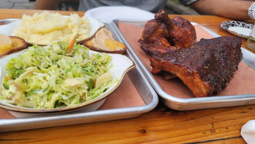 A plate with mashed potatoes, coleslaw, and grilled chicken alongside a tray of smoked ribs.
