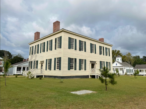 A large, two-story yellow house with green shutters, surrounded by grass and trees under a cloudy sky.