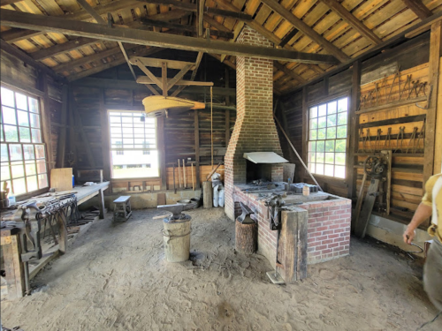A rustic workshop with wooden beams, a brick fireplace, tools on the walls, and a dirt floor. Sunlight streams through windows.