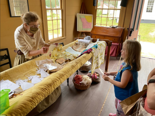A woman in period clothing demonstrates crafts to a young girl in a historic setting, surrounded by fabric and materials.