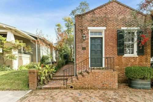 A charming brick house with a green door, surrounded by greenery and a brick pathway.