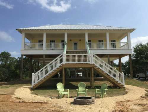 A two-story yellow house on stilts with a white staircase, surrounded by green chairs and a fire pit in the yard.