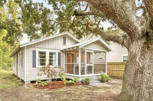 A charming single-story house with a porch, surrounded by greenery and a large tree in the front yard.