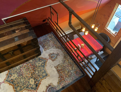 A cozy interior view from above, featuring a wooden railing, a patterned rug, a dining table, and warm lighting.