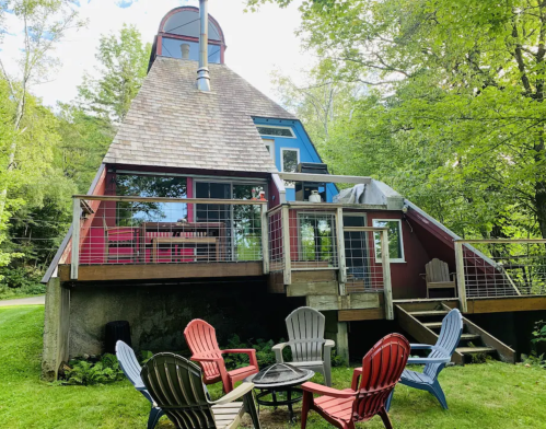 A colorful A-frame house surrounded by trees, featuring a deck and a fire pit with chairs in the foreground.