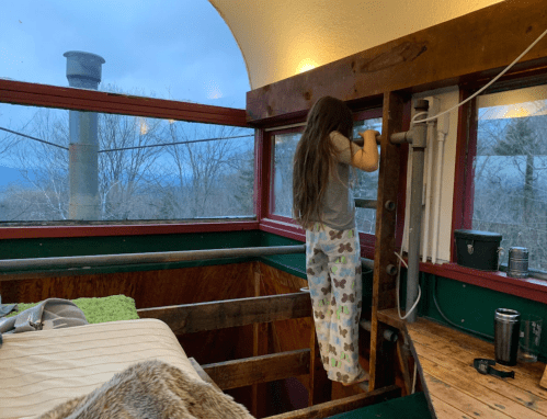 A child in pajamas climbs a wooden structure inside a cozy room with large windows and a scenic view outside.