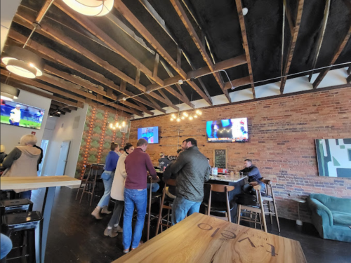 A lively bar scene with patrons socializing, exposed brick walls, and TVs showing sports in a cozy atmosphere.