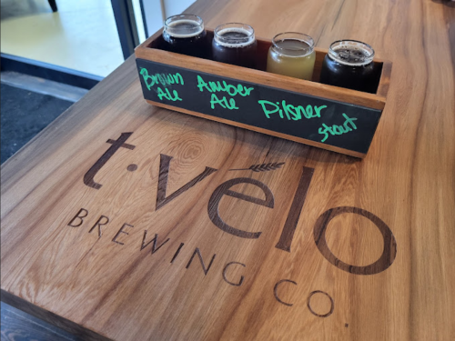 Three beer samples labeled Brown Ale, Amber Ale, and Pilsner on a wooden tray, with a brewery logo in the background.