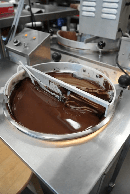A metal bowl filled with melted chocolate, with a mixing tool resting inside, in a kitchen setting.